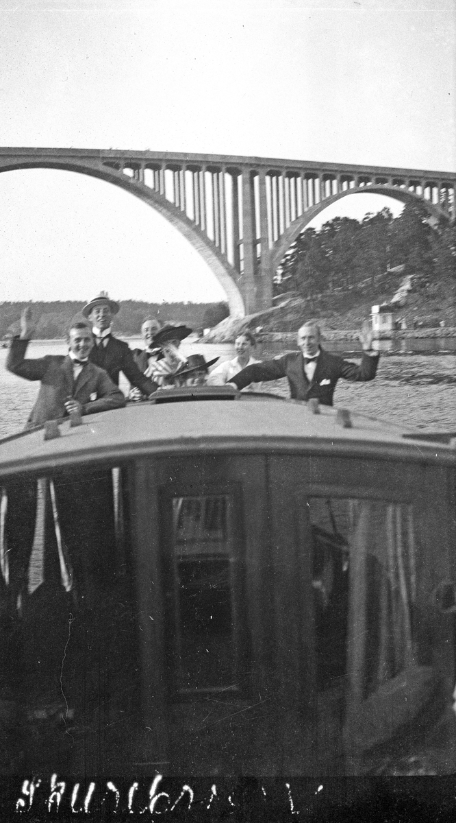 Gruppebilde. Fem menn og to kvinner står og vinker på dekk til en båt. Bro i bakgrunnen. Uleselig tekst nederst på foto. Hverdagsfoto.