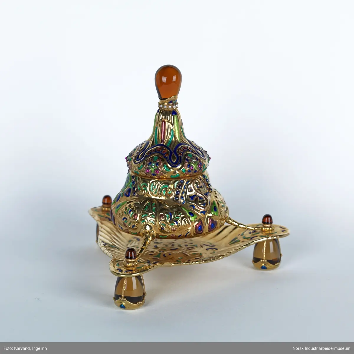 Blekkhus i gull - 750 (18 karat) - med rike former og sisselert. Står på tre ben og er dekorert med ulike stener som muligens er safirer, rubiner, smaragder, perler, citriner og sølv.

Dimensjon: 17 x  17 cm