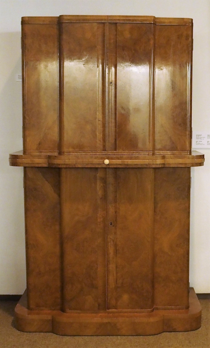 Cockailskap i to høyder med fire dører. Profilert korpus, sokkel, gurtlist og topplist. Mahogny med valnøttfiner; de øvre dørene er finert med lyst satintre på innsiden. Nedre skap har en hylle og en skuff festet øverst. Gurtlisten har en uttrekkbar hylle med ferskenfarget speilglass. Øvre del av korpus har dører som åpner til skap med belysning. Veggen bak, bunnen og to hyller er dekket med ferskenfarget speilglass. Øverst på veggen bak ses geometrisk dekor flankert av bladranker. På hver av dørenes innsider en hylle og gjennomhullet feste for cocktailpinner. Ti originale glasspinner med svaner er bevart.