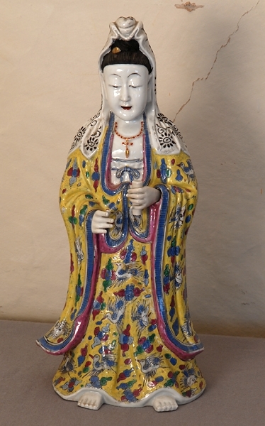 Ostindisk figurin av vitglaserat porslin föreställande stående person, vars långa klädnad och mantel är dekorerade med fåglar och geometriska figurer i blått, rött, grönt och vitt på gul botten med blå och röd kant på manteln. Händerna är rörliga och fästade vid handlederna med hjälp av två lösa ståltrådar som är dolda av manteln. Ena handen håller en rulle. Ögonen är halvslutna, munnen röd, pärlsmycke om halsen samt huvudklädnad i vitt med svarta mönster fastsatt på den bruna hårknuten.