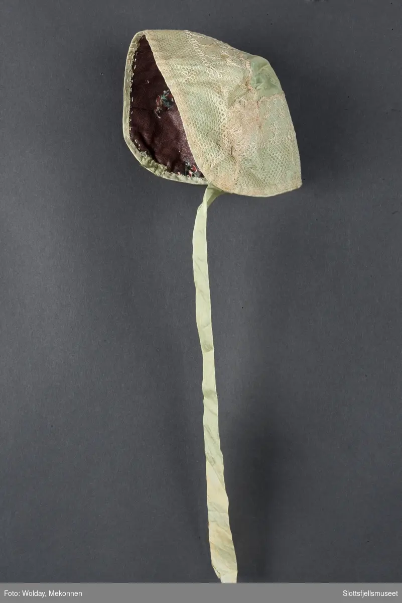 Dåpslue: av samme stoff som capen, sydd av et rett midtstykke og 2 avrundede sidestykker. Pyntet med bred silkeblonde, ecrufarget. Fóret m. Rødbrun blomstret mugg. Ett grønt silkeband, det andre mangler.