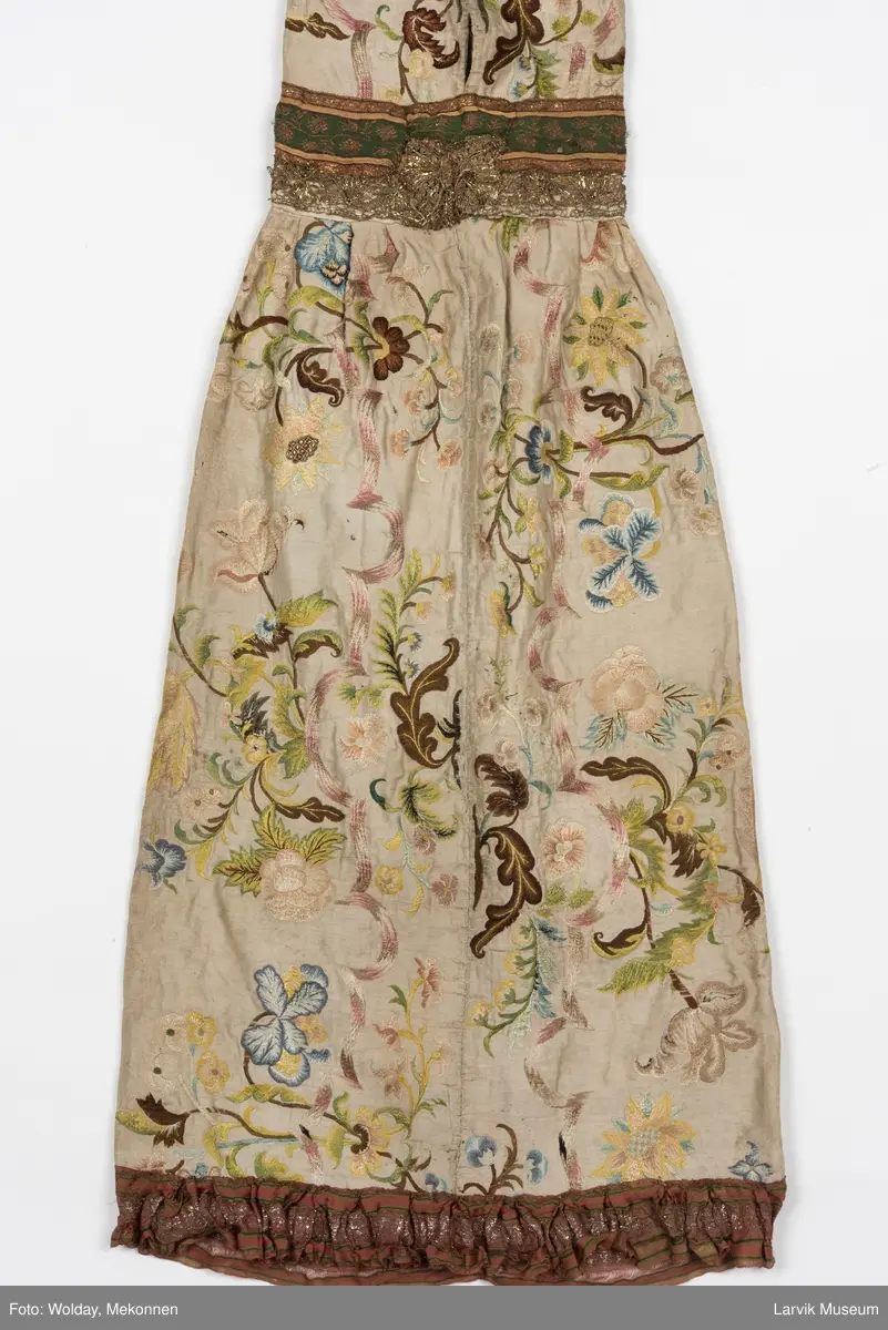 Form: livstykke som hette, åpen til livet, bred linning, rynket skjørt med blondekant nederst, søm bak på hetten i linning, søm foran og bak i skjørt, enkelte skjøtesømmer, linning brodert med gulltråd, skjørtet er foret med tynt stoff