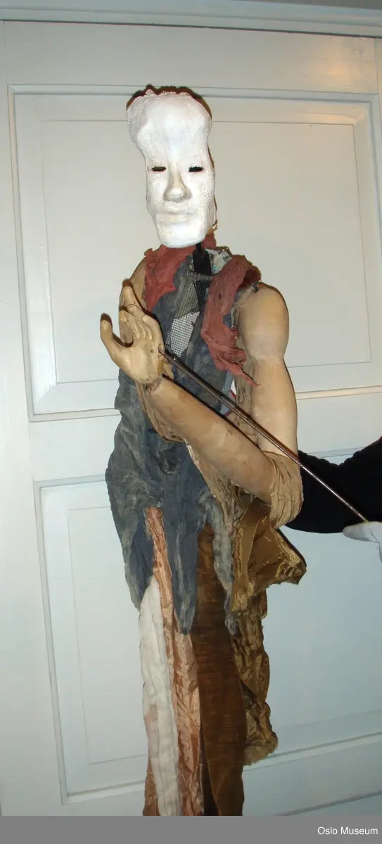 Stangdukke, mann fra "Antigone" av Sofokles.
Ansiktet er gips og gasbind. Håret er stål som er bøyet og formet med strimler av gasbind og gips rundt om på hodet. Inne i masken er det sort plast og teip. Hodet står på en trestang som er festet med plasttråd. Trestangen går ned gjennom brystet som dukkeføreren kan bruke til å bevege hodet med.Trestangen  er festet med stål og hemper.
Brystkassen er hønsenetting. Farget gasbind i blå og rosa er limt på. Armene er i tre og bevegelig i skulder-, albue- og håndledd. Skinnlapper mellom treverket danner leddene.
Dukken har på seg forskjellige tøystoffer som henger løst ned langs kroppen i blå, fersken og brun. Armene er festet til hønsenettingen med et beige silkeaktig stoff som henger ned langs armene.