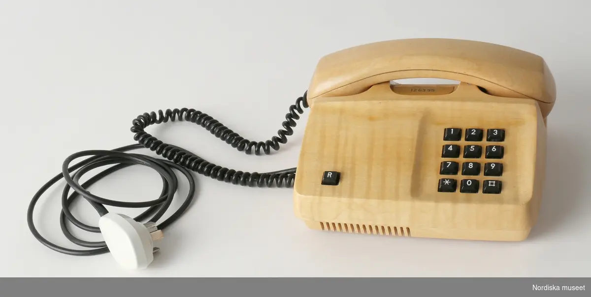 Telefon, för fast telefoni, med hölje av lackerat trä (Sykomorlönn). Knappsats med svarta plastknappar med siffrorna 1-9 samt stjärn- och fyrkantsknapp. Även R-knapp till höger. Svagt svängd lur med mikrofon och högtalare i var ände. Mellan luren och telefonapparaten löper en svart spiralformad sladd av plast innehållande telefonledning. Luren vilar i urtag i telefon. Vid urtaget är fäst en mässingsbricka med telefonnummer "12 63 99" (Kommentar från konservator: möjligen har mässingsskylten en skyddslack eller eventuellt ett guldskikt som skyddar från oxidering). Undertill har telefonen en bottenplatta av plywood med fyra gummifötter, vitt reglage för ljudnivå samt text i guldfärg inom fyrkant: "Teli. /ART.NR DBAA 103 905/4 / TILLHÖR TELEVERKET / CERTIFIKAT No 40 / SYKOMORLÖNN  / TILLV. ÅR 1981.".
Från höger sida av telefonapparaten en svart plastklädd sladd, som avslutas i vit telefonkontakt (att fästas i telefonjacket) med metallpiggar. Innanmäte med div. elektronik.
Anm. Ett urslag vid urtaget för luren.
/Leif Wallin 2017-04-06