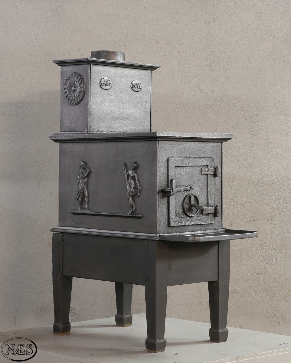 Kokeovn med to muser på enkel profillist. En fløytespillende og en dansende. Begge med draperier. Kokeanretning i topplaten til 1.etg. Ovnsdør på kortside. Rosett på begge sideplatene 2.etg. Den ene er løs og fungerer som ventil. Innskrift på frontplate og bakplate 2.etg. Røykuttak i toppen.