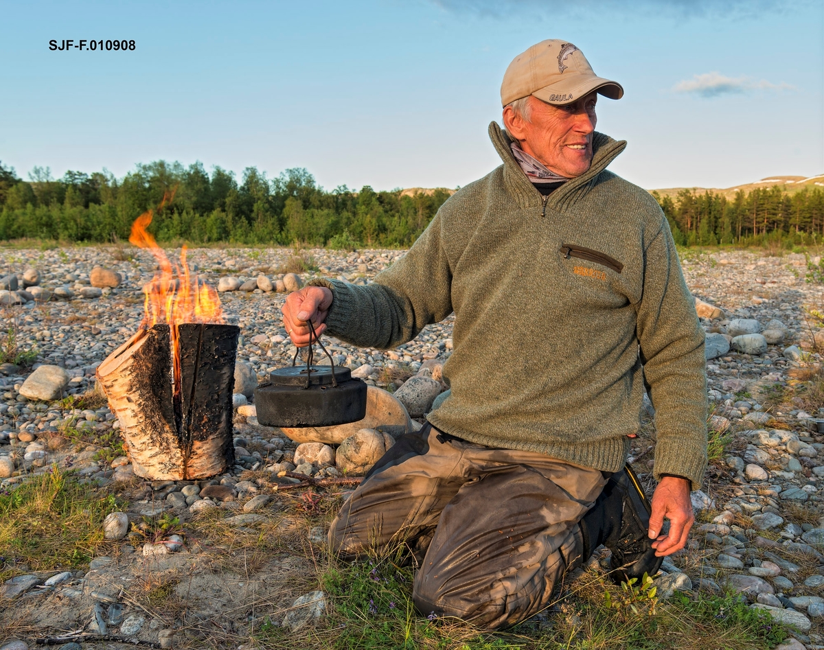Idar Edvardsen fra Bjerkvik (opprinnelig fra Grovfjord) med svartkjele. I bakgrunnen en finneprimus eller stubbfyring som han har brukt til kaffekoking. Friluftsliv. Leirliv. Kokekaffe. Kaffe. Bildet er tatt ved Altaelva i Øvre Alta, Finnmark. Idar har nettopp landet en laks på 15 kilo i Altaelva, og det var god stemning i leiren. Laksefiske i Altaelva, Alta, Finnmark. Fiske etter laks. Sportsfiske.