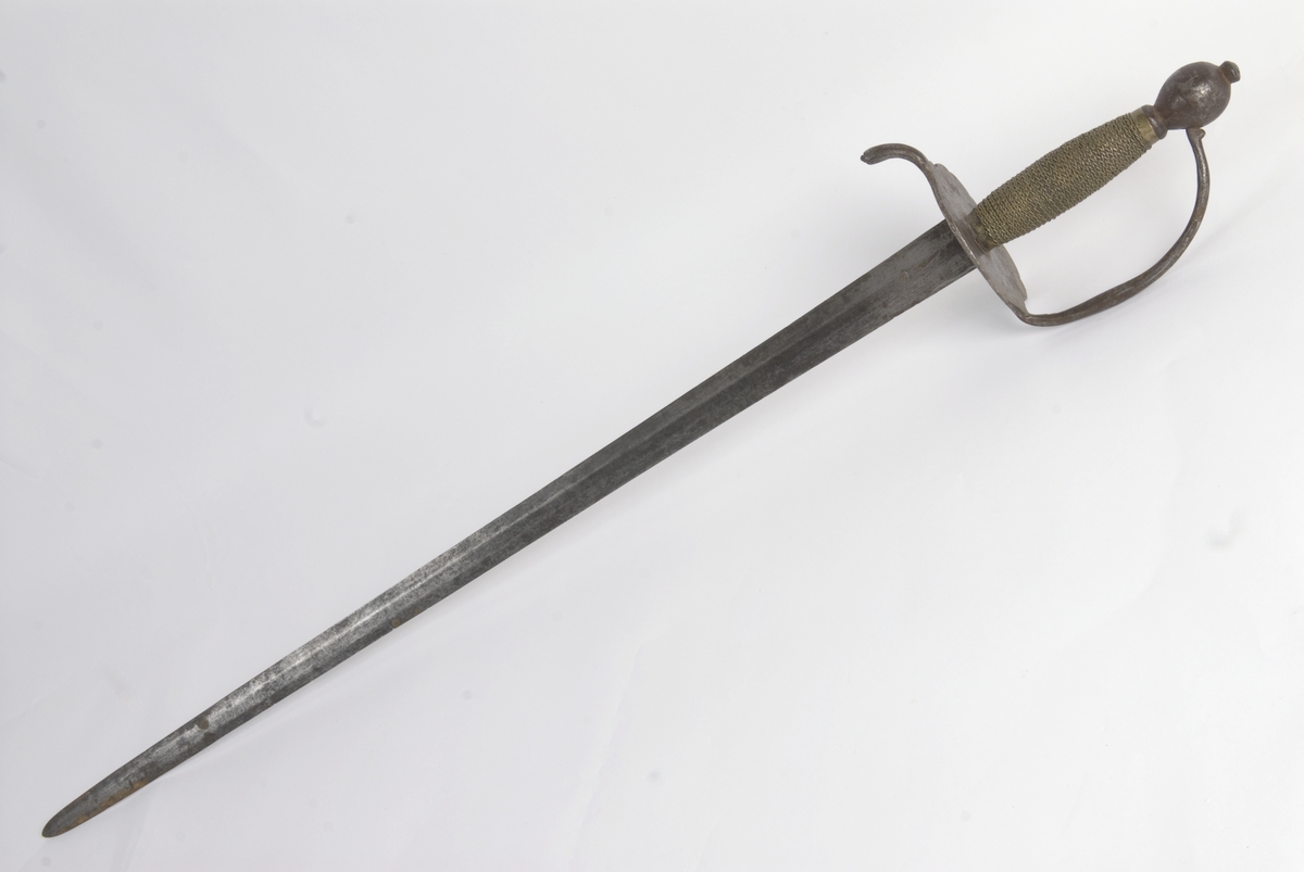 Dansk/Norsk Soldatkårde modell 1701. Plommeformet knapp med nitteknapp. Tregrep med messingtråds bevikling og hylse oppe og nede. Nyreformet parerplate med 5 fliker. Nedbøyd bakre parerstang. Fremre parerstang går over i håndbøylen og er tappet i knappen.
Rett tveegget klinge med 6 kantet tverrsnitt. Rester av etset dekor.