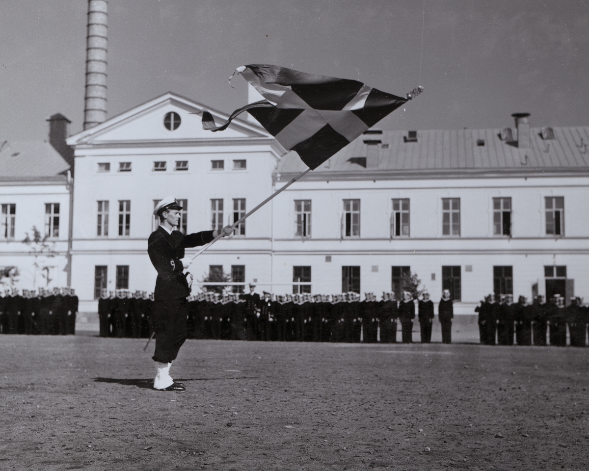 En uniformsklädd fänrik står på Sparres kaserngård med Sveriges flagga i händerna. I bakgrunden syns uppradade flottister framför matinrättningsbyggnaden.