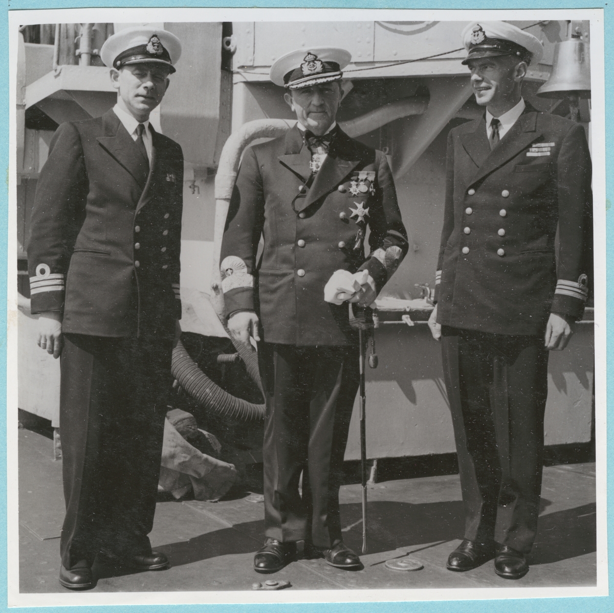Konteramiral Samuelson poserar framför kameran ombord på ett fartyg tillsammans med två kaptener. I ena handen håller han en sabel och en handske. Från Danska och Norska örlogsbesöken den 29/6 - 4/7 1951.
