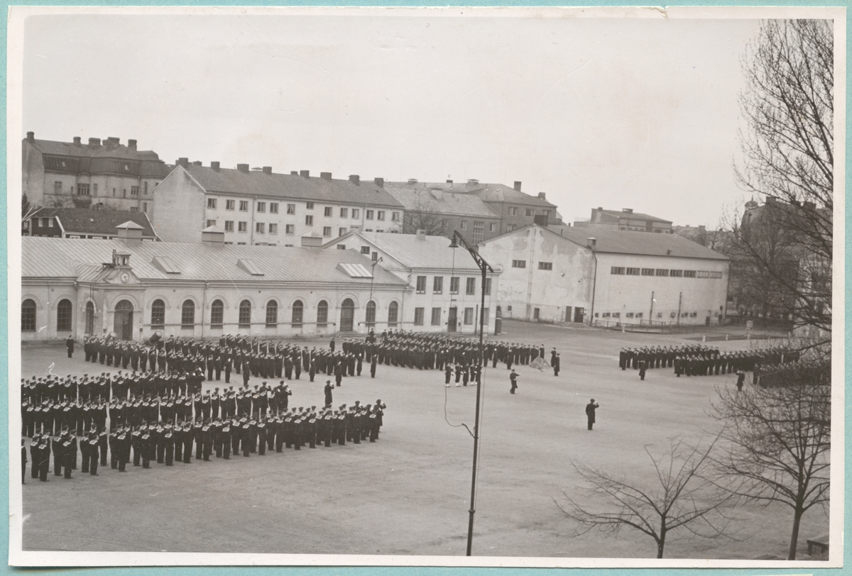 Grupper med flottister i uniform står på led inne på bataljon Sparres kaserngård. I mitten står befäl och högre militärer. Runt omkring grupperna syns bataljonens excercisbyggnad samt simhall.