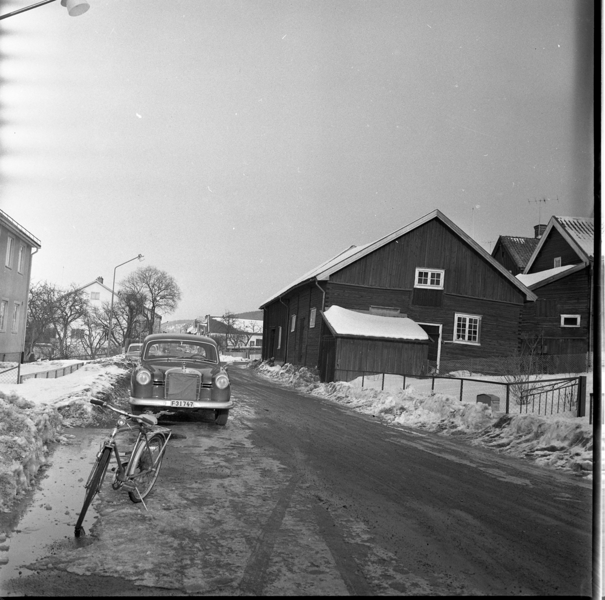 Vinterbild med Sjögatan norrut. En cykel och en Mercedes Benz (F31747) parkerad mot kameran och till höger ekonomibyggnaden till Hallska gården.