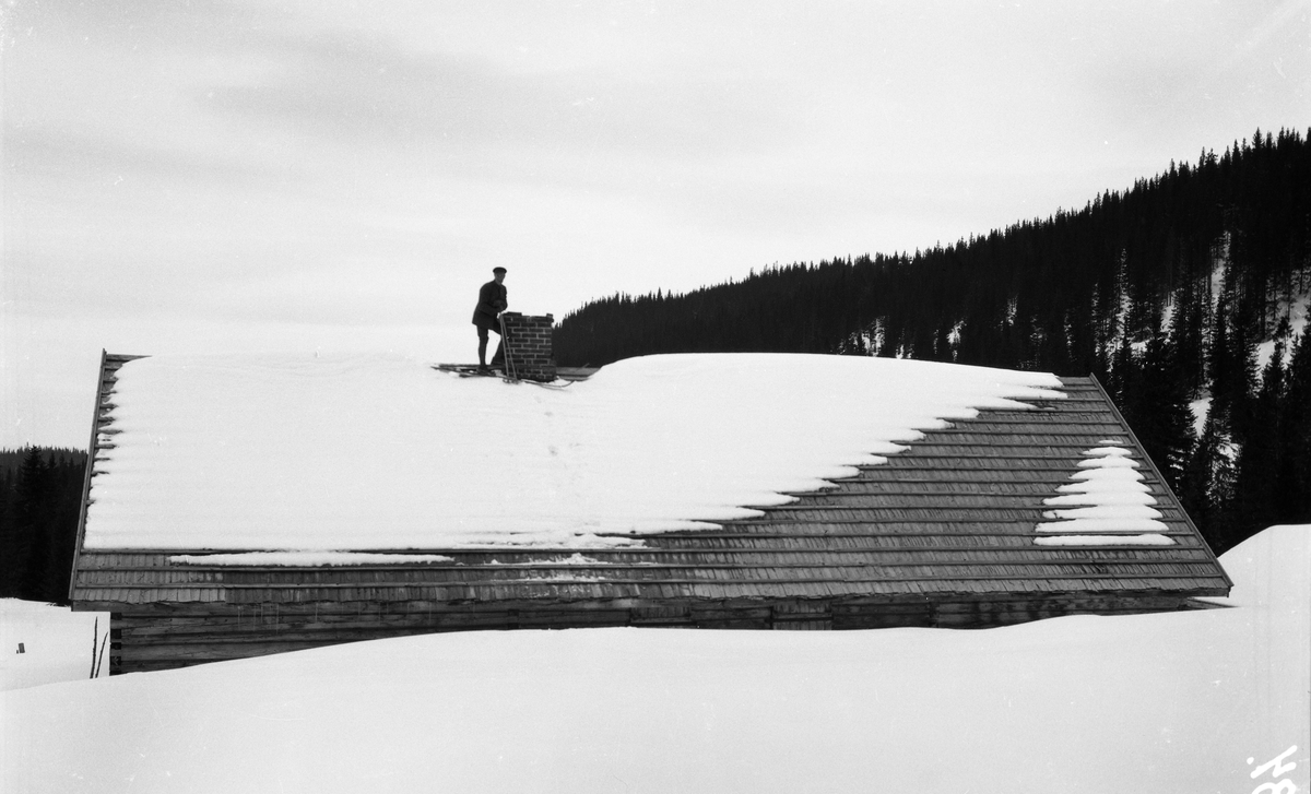 Snøvinter. En skiløper står på taket av noe som trolig er et seterstørhus. Hverken person eller sted er identifisert.
