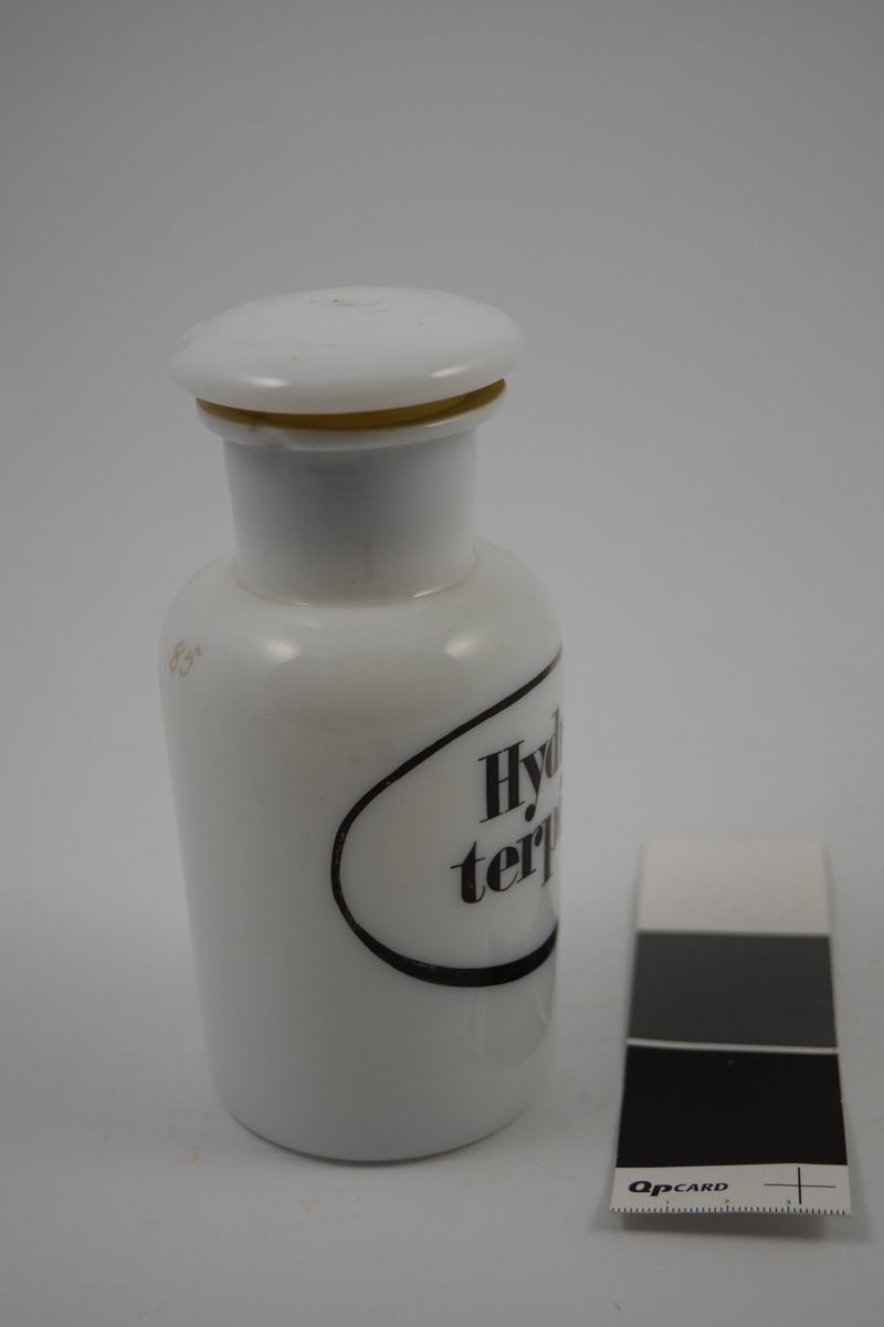Porselenskrukke med propp. Sort oval etikett påført på krukken. Til oppbevaring av legemidler/pulver.