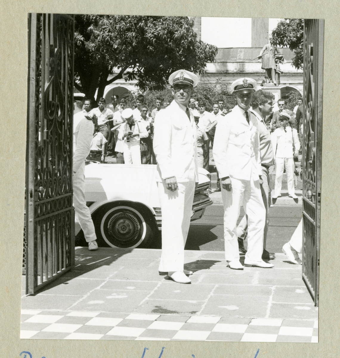 Bilden föreställer två män i vita uniformer som står i en grindöppning. I bakgrunden syns en bil och ytterligare vitklädda personer. Bilden är tagen i Puntarenas under minfartyget Älvsnabbens långresa 1966-1967.