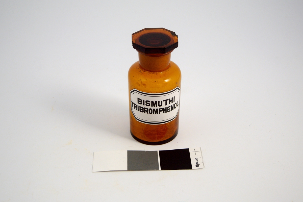 Brun glasskrukke med glasspropp. Hvit etikett med sort skrift. Bismuthi Tribromphenol er et desinfiserende middel.