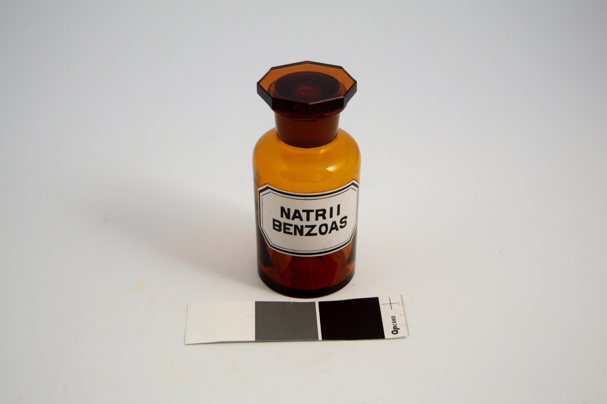 Brun glasskrukke med glasspropp. Hvit etikett med sort skrift. Natrii Benzoas ble brukt som konserveringsmiddel i diverse legemidler.