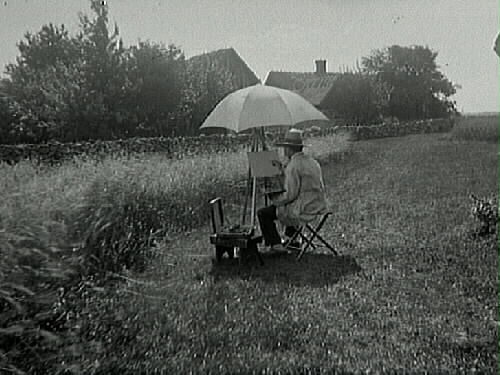 Konstnär vid staffli med parasoll framför en gård med halmtak och stengärdsgård.