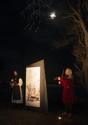 Munk i hvitt og svart synger mens mann i rød middelalderjakke og struthette spiller fiolin. Over lyser månen fra den mørke høsthimmelen.