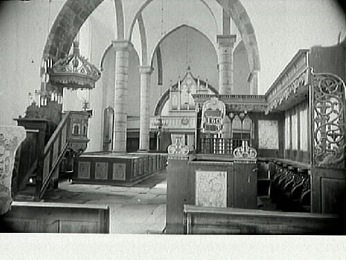 Interiörbild av okänd kyrka, rikt dekorerad. Vy över långskeppet med slanka pelare mot orgelläktaren. Till vänster i bild predikstol med genombruten baldakin och till höger korbänkar och psalmnummertavla.