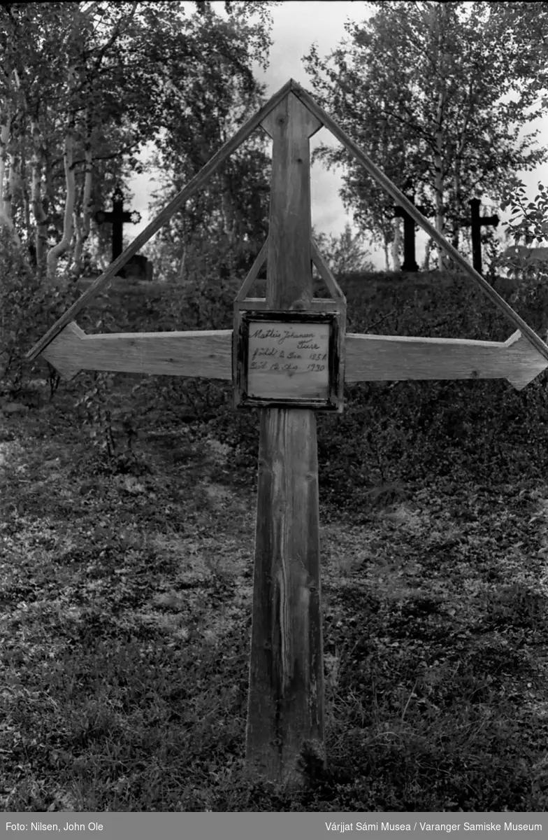 Gravkors av tre, sannsynligvis utenfor Kautokeino kirke. Gravkors av metall og trær i bakgrunnen. Juli 1967.
