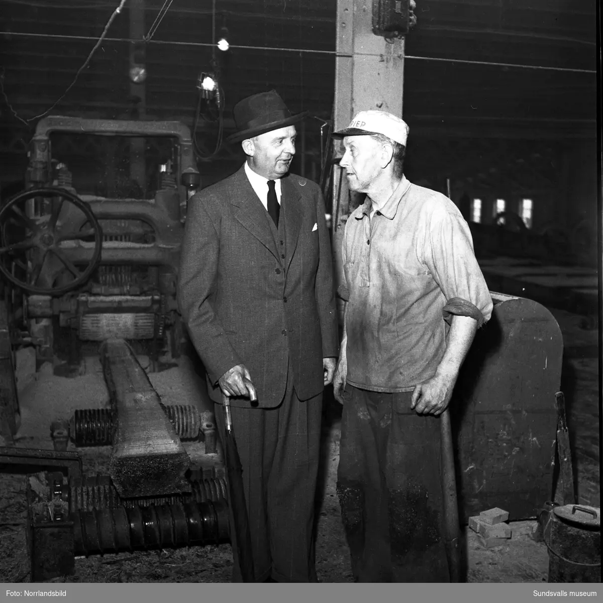 En man i hatt, kostym och slips pratar med en arbetare i såghuset på Skönviks sågverk.