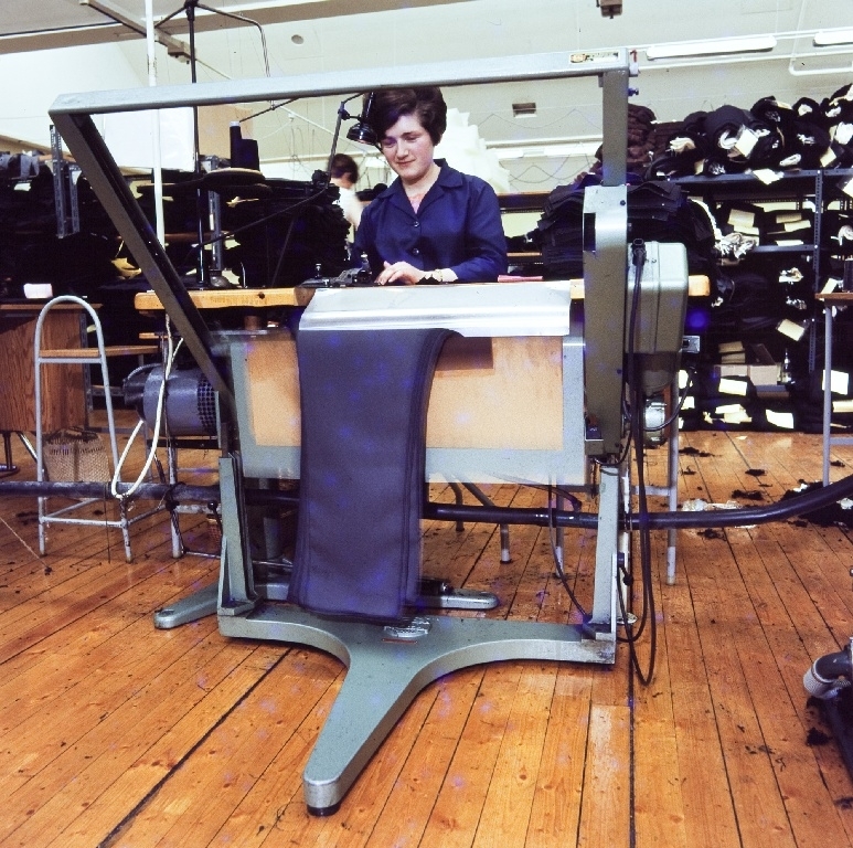 Kvinnelig operatør arbeider med overkasting av sømmer i bukser med automatisk trådkutter i konfeksjonsfabrikken til Jonas Øglænd Sandnes. Bak stables de ferdige delene etter sying.