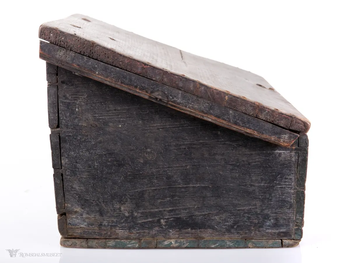 Rektangulær skrivekasse med skråstilt lokk, hengsler og låsbeslag. Bunnen er delvis dekket av et tykt lag med stivnet blekk med en trepinne i.