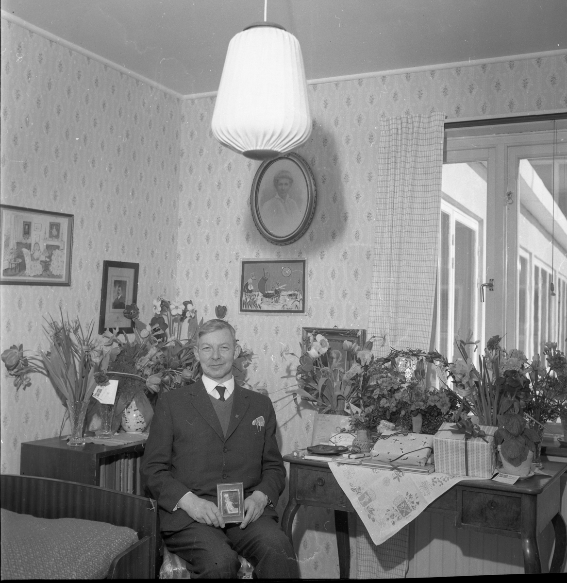 Louis Stackelberg fyller 50 år, mars 1964. Sitter på en stol omgiven av blomsterbuketter med en liten ramad tavla i händerna.