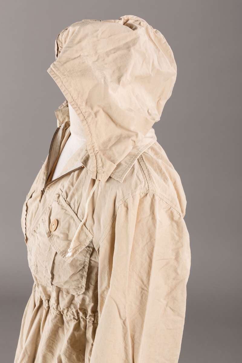 Form: Vanlig anorakkfasong med strikk i livet. 2 brystlommer med knapp.
Hette med glidelås på topp midt, snøring.
Glidelås helt ned foran.
Innermansjett med strikk i ermene.