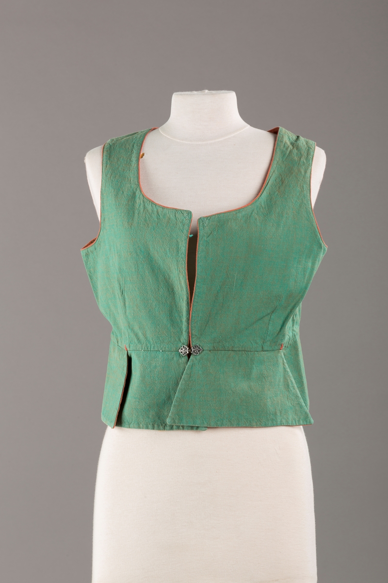 Grønt liv med rustrød kant og fór, seks skjøter. Håndsydd forsterkning i kontrastfarge øverst i skjøtesplittene. Lukkes med tinnhekte.