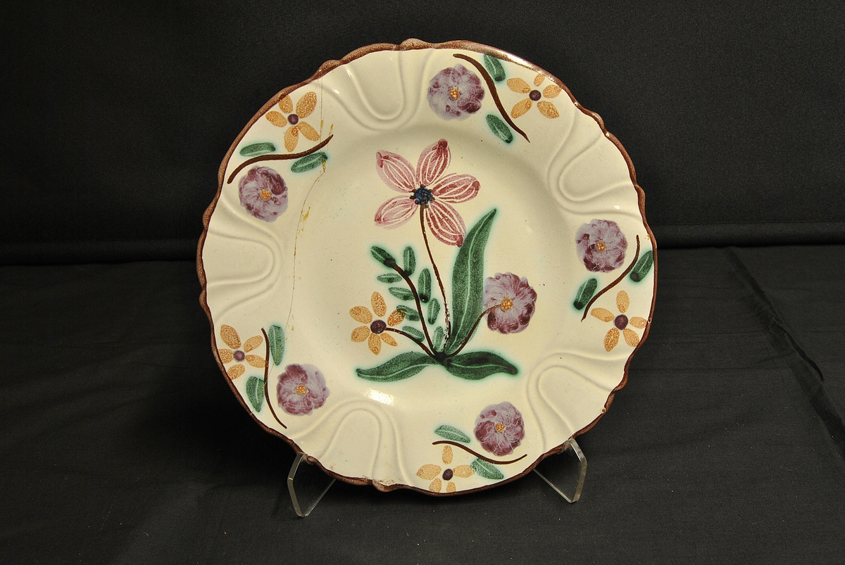 Sirkelforma, kvit tallerken med bølga kant og blomsterdekor: eit stort motiv på midten og fem mindre motiv rundt kanten.
