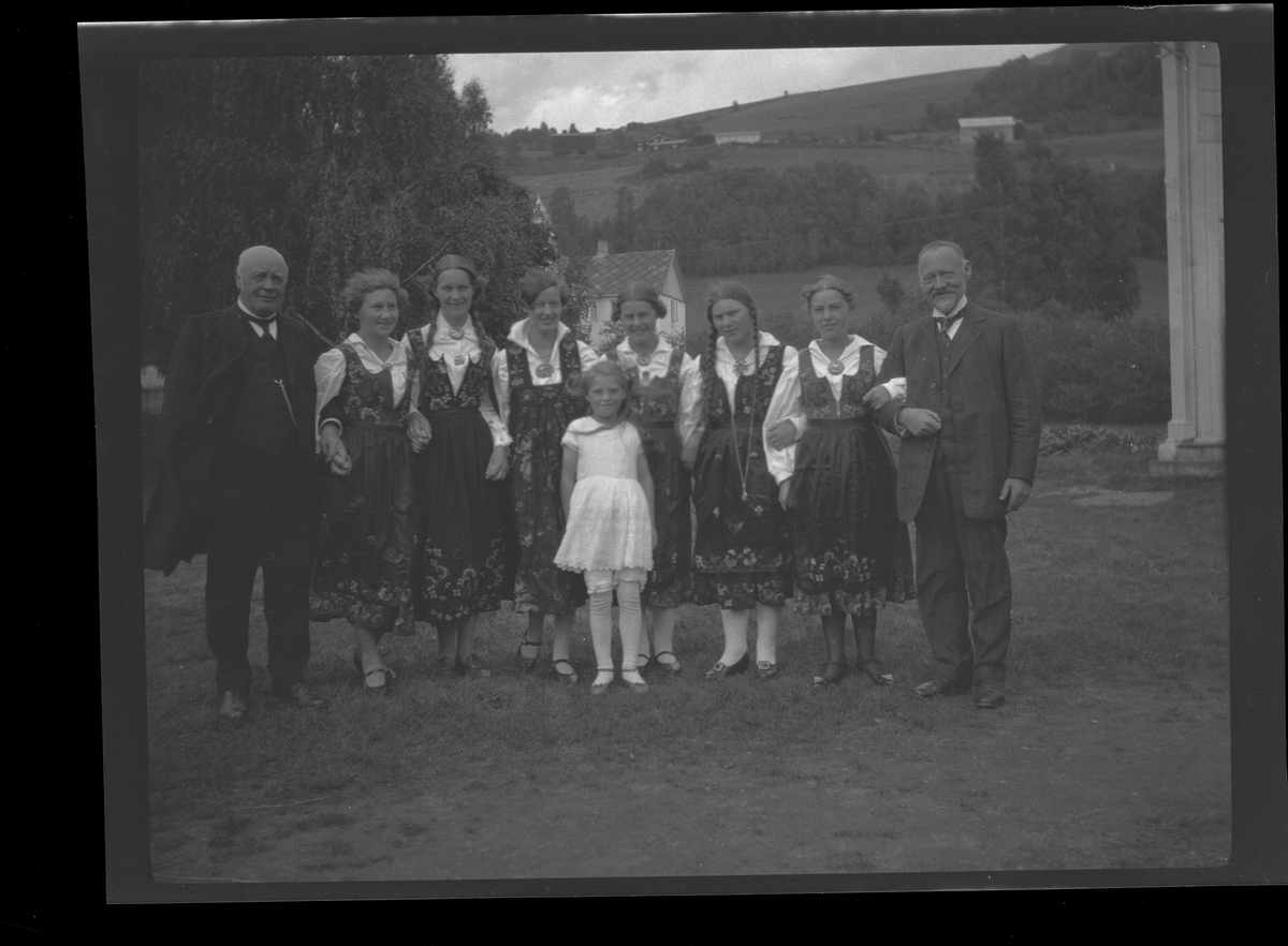 Rasmus Stauri til venstre med 6 bunadskledde kvinner og en eldre mann til høyre. En ung jente i front. De står ute på tunet.