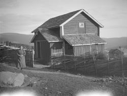 Eldre dame står forran 2 etg. hus med skifertak.Huset ligger