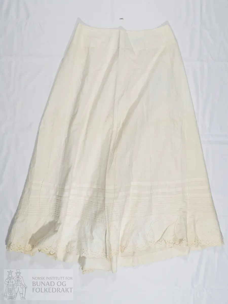 Understakk i hvitt bomullslerret. Bred linning som lukkes med en trukket hvit knapp. Linningen har en skjøt. Flere legg nede på stakkekanten, to bredere og flere mindre. Helt nederst er stakken påsydd en blondekappe, så nederst består stakken av to lag stoff. Blonden er av bomull med engelsk broderi. Stakken er sydd sammen av 5 høyder. Kun maskinsøm.