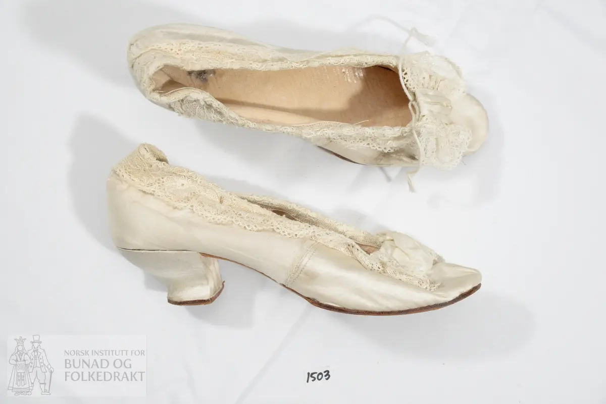 Sko trukket med hvit silkesateng. Påsydd kjøpeblonde i bomull langs åpning, sløyfe av blonde og silke på overlæret. Såle av skinn. Skinn i selve skoen. Snellehæl.