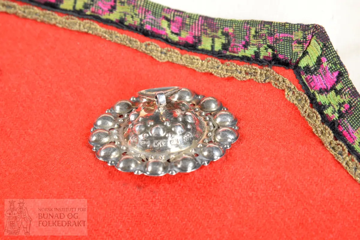 Liv sauma av raud ullkypert, lett valka. Fora med ubleika bomullslerret.
Ermhole, rundt hals og opning framme er kanta med blomstermønstra fleirfarga fløyelsband med svært avslitt fløyel. Innanfor kantingar er det påsett eit smalt metallband.
Etter framkantane er det påsett 3 borer på kvar side, dei to øverste litt breiare enn dei fire nederste, alle har eit skåleforma lauv festa i midten. På framsida av den kurva delen er det fem ulike stempel: 63 i eit sirkelforma stempel, truleg årstalsstempel. Månadsstempel: 6 over M i rektangelforma stempel. Mesterstempel i bølgeforma ramme LAR, Lorentz Christopher Angel Reimers, fekk borgarskap i Bergen i 1832. Lødighetsstempel 13 1/3. Trapesforma stempel med 7 prikkar, Bergens bystempel.
Lukking framme med to metallhekter på kvar side.
Heile livet er sauma på maskin. Kanting er sauma for hand. 
Livet sauma på stakken for hand. 
Stakken er av heimevove ullerret, blåsvart. Splittopning midt framme. Samansett av 5 rette høgder med jare i begge sider, alle høgder. Faldelagt oppe med eit glatt felt framme mot splitt. Høgdene er sauma saman med maskin. 
Nedkant: 3 svarte smale sillkefløyelsband, nedanfor dei eit påsett stoffstykke av svart bomullsfløyel. Dette stoffet er ein hengefald, ikkje sauma fast i nedkant.
Nedkanten er skonna på innsida med eit lerretsvove mohairstoff av mørk grøn farge. Sauma på med maskin. I nedkant av skonninga er det påsett eit blåsvart kosteband sauma på for hand.
Kanten på linninga er av svart bomullssateng. Er bretta dobbelt og sauma på for hand på begge sider