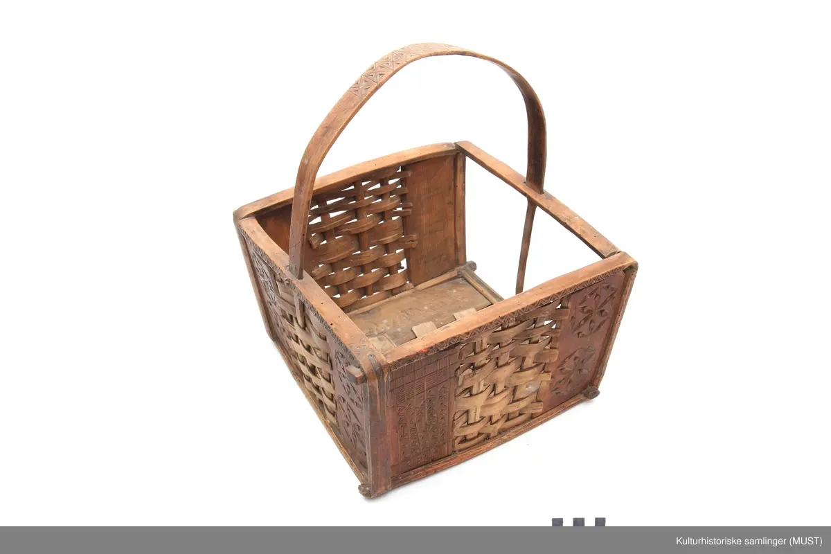 Firkantet trekurv med flettedekor og treskjæring på både kurv og håndtak. Dekoren består av to seksbladete blomster på hver side samt ulike trekantmønstre langs kanten av kurven og håndtaket. En av sidene i kurven mangler.