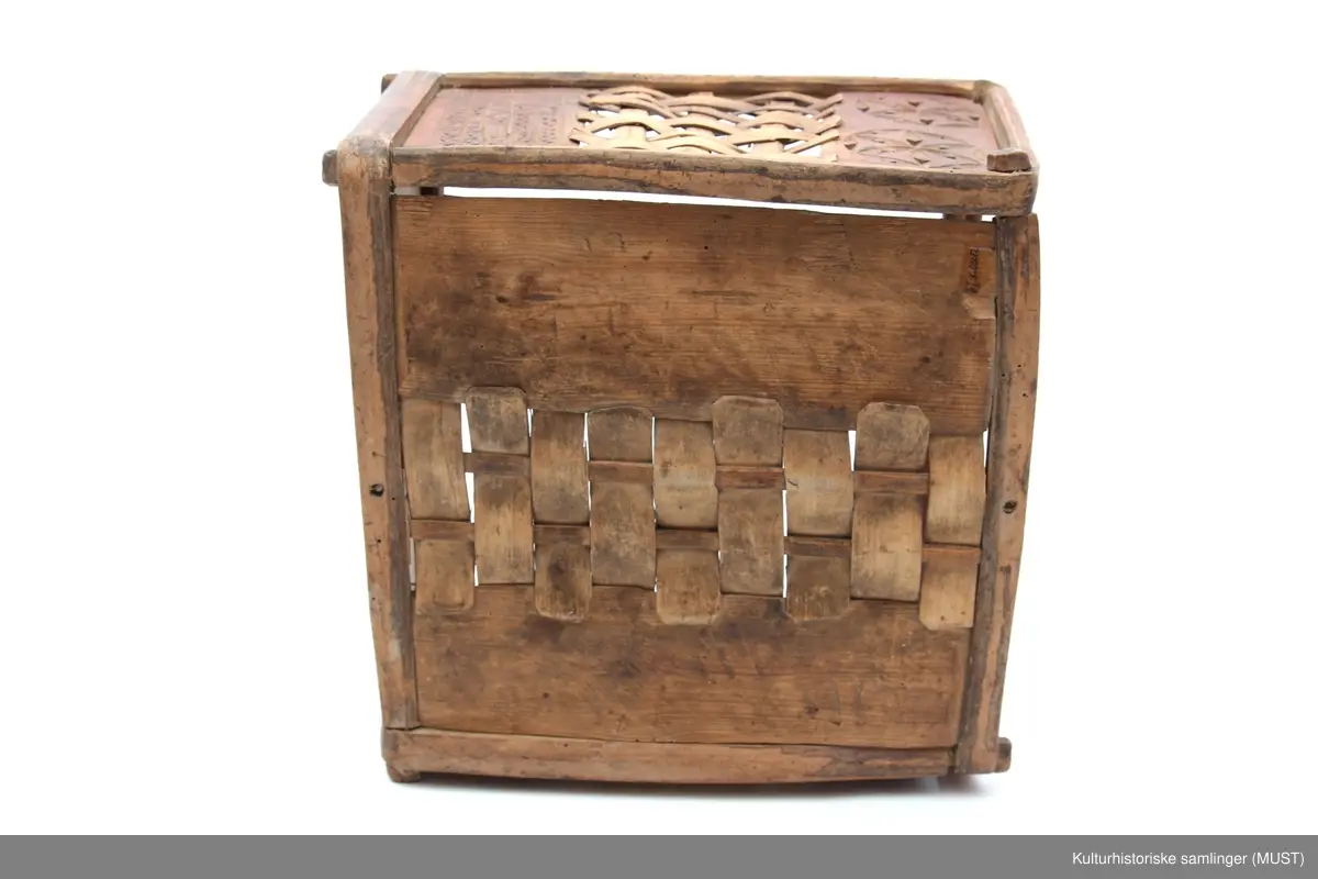 Firkantet trekurv med flettedekor og treskjæring på både kurv og håndtak. Dekoren består av to seksbladete blomster på hver side samt ulike trekantmønstre langs kanten av kurven og håndtaket. En av sidene i kurven mangler.