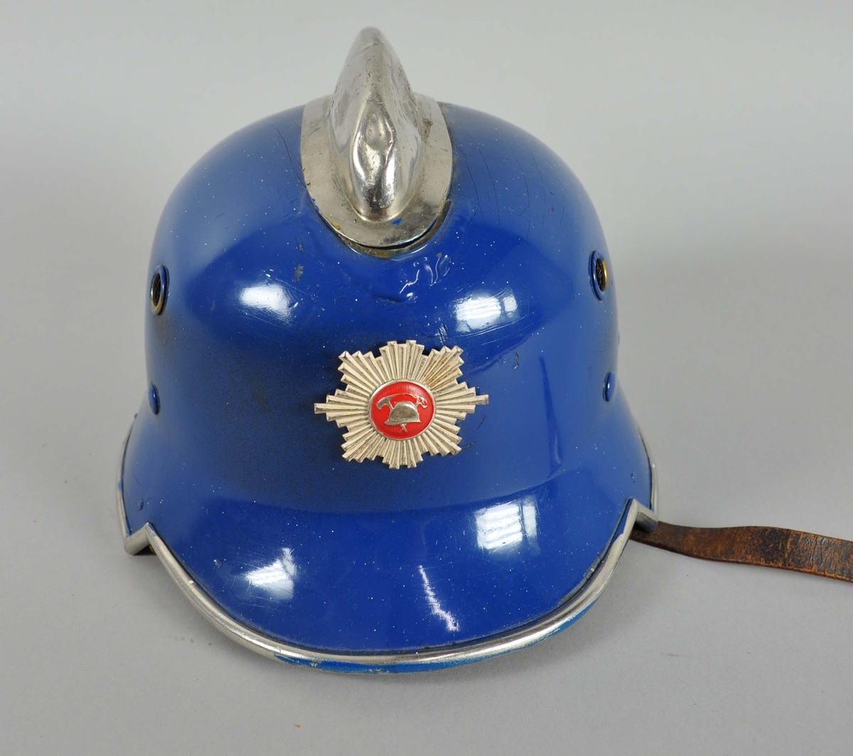 Blå brannhjelm av metall. Hjelmen har innerhjelm og hakereim av lær. På toppen er går det en opphøyd kant eller hanekam. På framsiden er emblem for det norske brannvesenet.