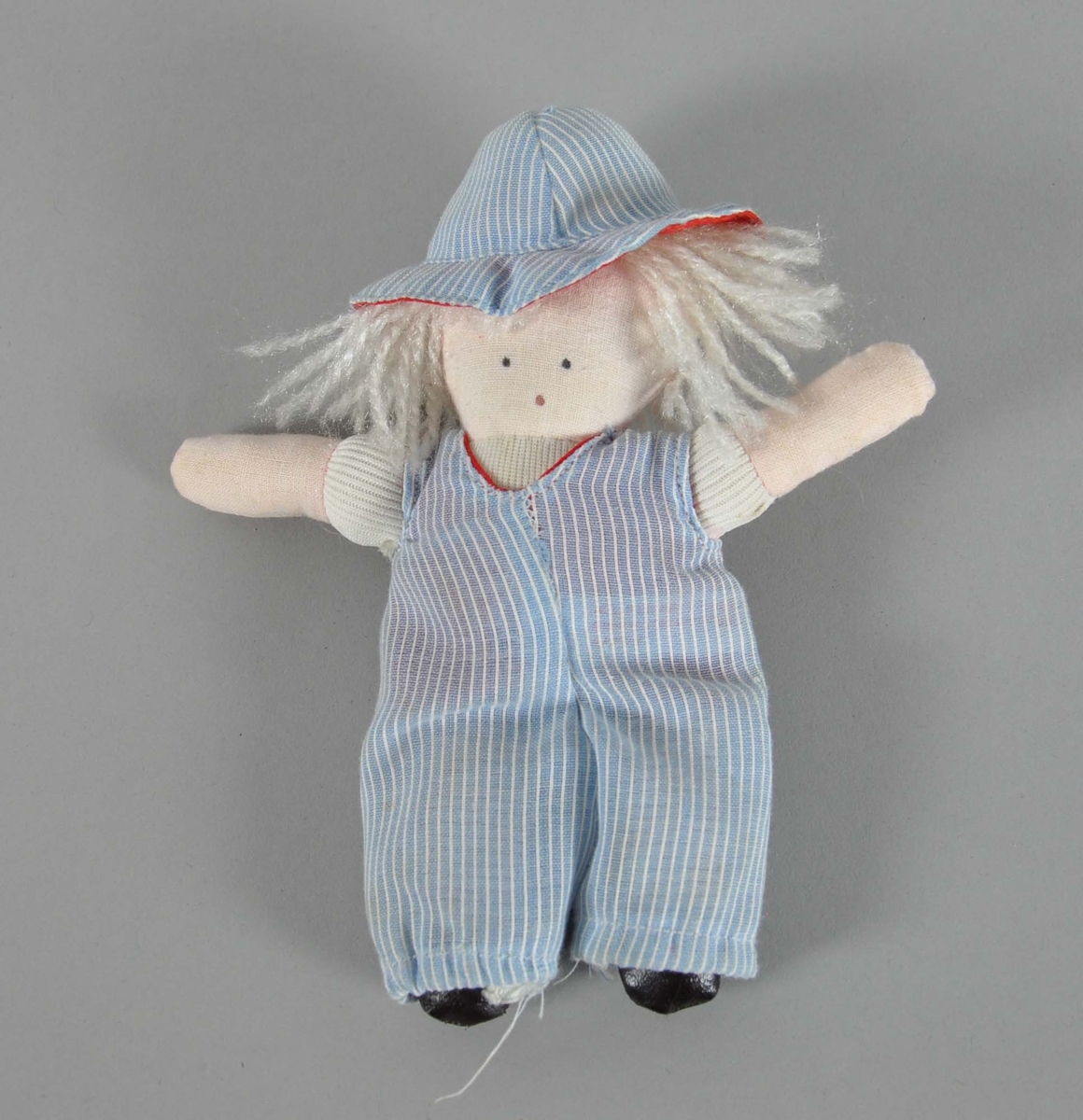 Hjemmelaget dukke. Dukken er hvit og har hvitt langt hår. Hatt og overall er blå med hvite striper.