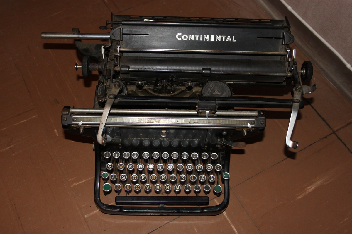 Rektangulær, mekanisk sort skrivemaskin med 49 tangenter på fire bein. Tangentene har sort bakgrunnsfarge.