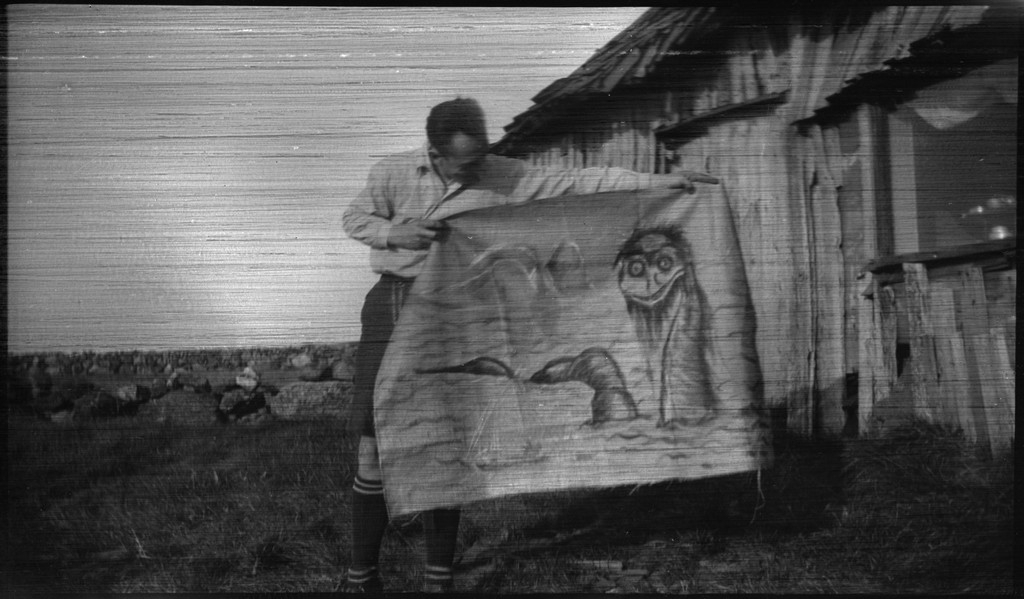 Lindtner, Otto Olsen og en til i og utenfor bua "Inmaribu" på Gauselholmen utenfor Stavanger. De har reist en bjørkestamme med tresopp på ved et steingjerde. Johannessen holder frem et maleri på tekstil av en sjøorm.