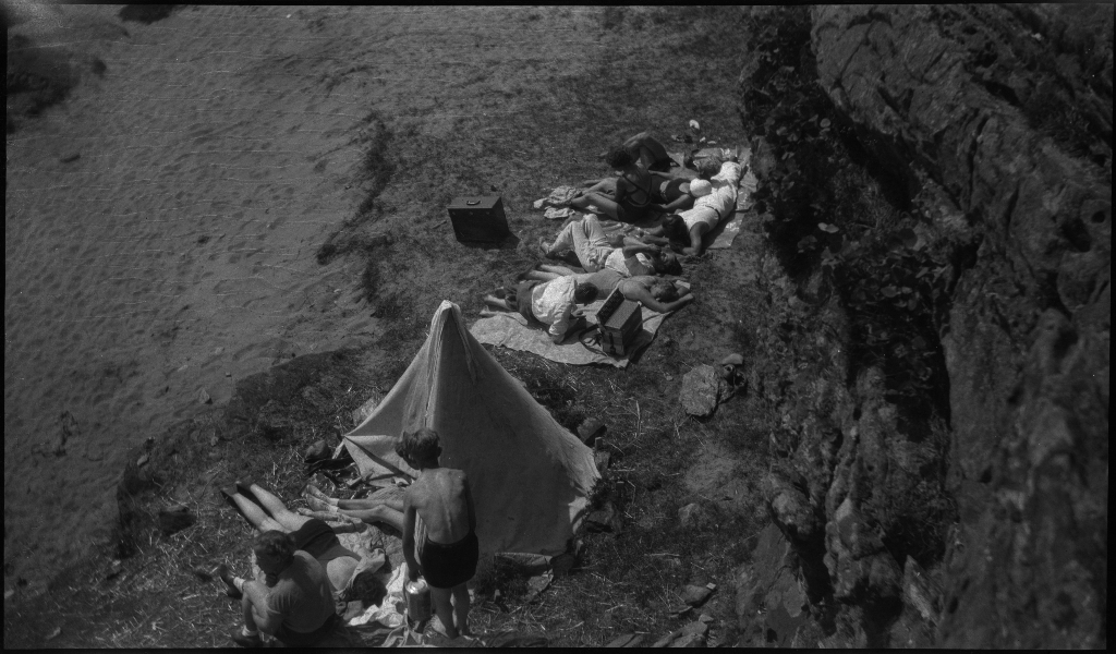 Barn og unge på og rundt stranda i Vigdelsvika. De fleste har satt opp telt, noen soler seg og noen bader. Den ene gutten på bilde nr. 6 holder et flytebelte.