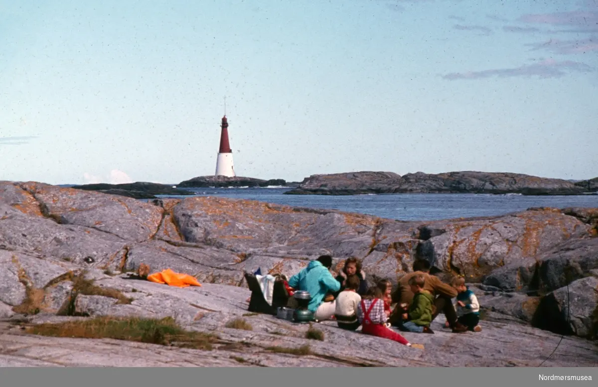 Foto av en familie på utflukt på fiskeværet Grip i Kristiansund kommune, med fyrtårnet i bakgrunnen. Fotograf er Kjell Haug og er den som står bak samlingen med fargefoto/dias fra Kristiansund og Grip. Bildene dateres til omkring 1950-1970. Fra Nordmøre museums fotosamlinger.