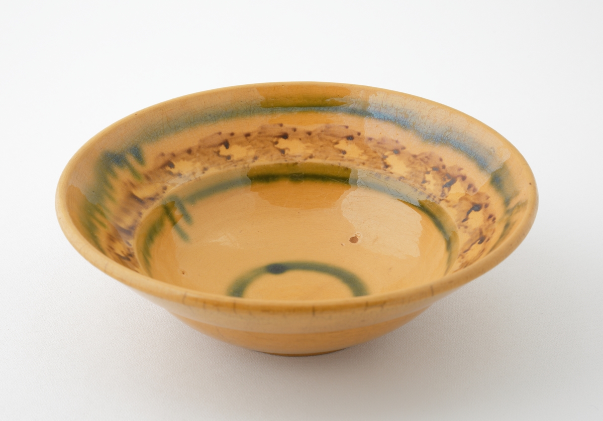 Skål i glaserad keramik.
Rund skål med honungsgul glasyr, försedd med invändig dekorrand (fjäderformationer) i brunt och två gröna ränder i rinningsglasyr. I botten en cirkelrund grön rand.