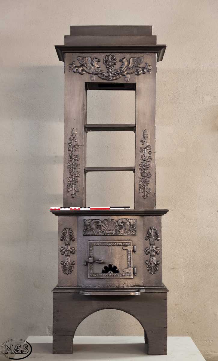 Ovnen består av to avdelinger. Underdelen har et frembuet midtparti med kamskjell flankert av sjødyr på hver side. På kortsidene skjold, løvehode og symmetriske ørner. På bakplate en krukke med symmetriske engler.
Overdelen er høy, firkantet med tredelt åpning. Empire ornamentikk og avtrappet topp. Øverst på frontplaten symetriske fabeldyr (pegasus) vendt mot hverandre.