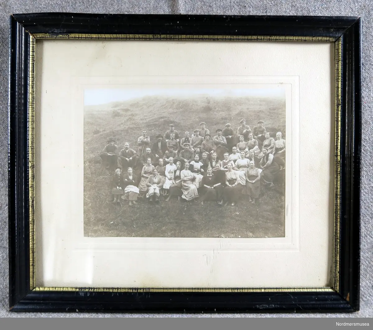 Bilde med svart ramme. Viser arbeidarane ved ullvarefabrikken i 1920 (?). Bilde er tatt på en grasbakke utanfor fabrikken.