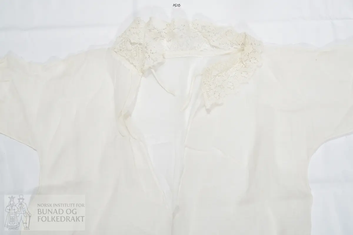 Dåpskjole i tynn hvit bomull, vevd i litt glissen lerretsbinding. Splitt midt bak, lukkes med knytebånd i nakken. Knytebåndet ligger i løpegang i halsen. Løpegang med knytebånd også nederst på ermene. Kragen er laget av kjøpeblonde/maskinprodusert blonde, samme kjøpeblonde er brukt som kanting nederst på skjørtet og nederst på ermene. Blondene er blitt stivet. Ikke tydelig definert liv, sydd med en lang sidesøm. Sidesømmen er sydd i ett med ermesømmen. Maskinsøm med håndsydde detaljer.