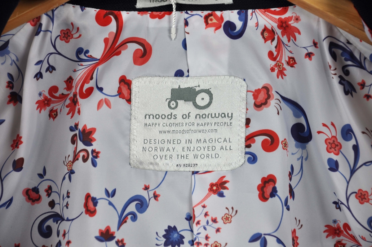 Mørkeblå dressjakke av ull/silke, foret med samme silkestoff som i damekjolen og tørkleet, et
hvitt silkestoff med "rosemalt" mønster.Jakken har fire innerlommer, en med knappelukking.
Utvendig to jakkelommer med klaff foret med silkestoffet, en brystlomme med påbrodert
"traktorlogo", Mørkeblått kantebånd fra kraven og ned. Lukket med en stor stoffknapp på ermene, fire ganger to mindre stoffknapper i hvitt (2), rødt og blått. På høyre erme brodert:Heia Norge i henholdsvis rødt og hvitt.
Str.:48