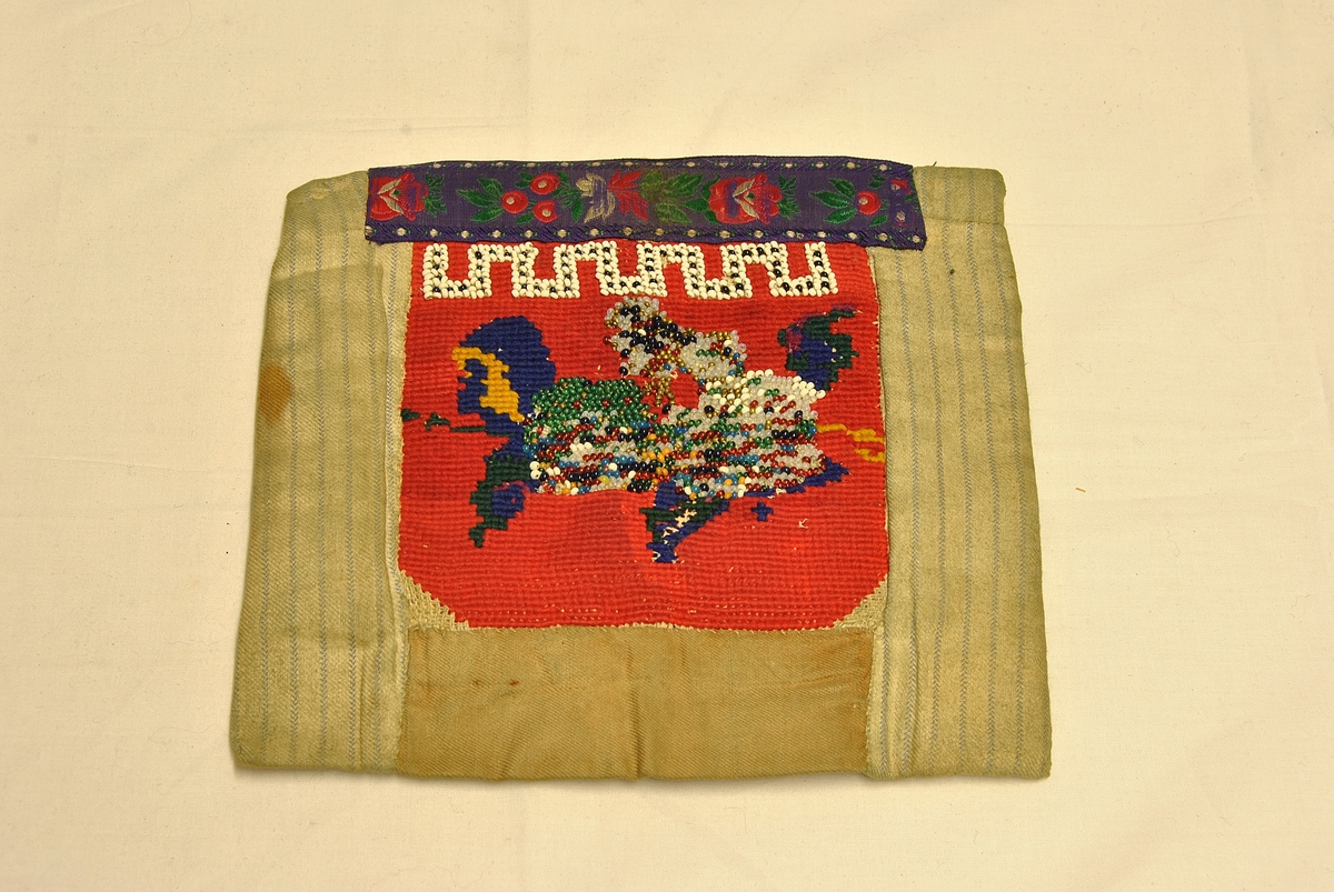 Nær kvadratisk form. Sydd sammen av flere tøystykker. Broderi i front, med glassperler. Pyntebånd oppe. Avstivet med papp (?) og tykt ullstoff inni.